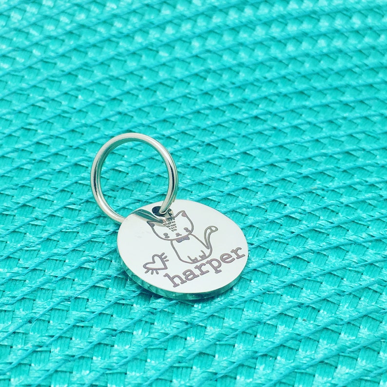 May include: A round, silver pet tag with a cat illustration, a heart, and the name "harper" engraved in a script font. The tag is attached to a silver ring. The background is a textured, turquoise surface.
