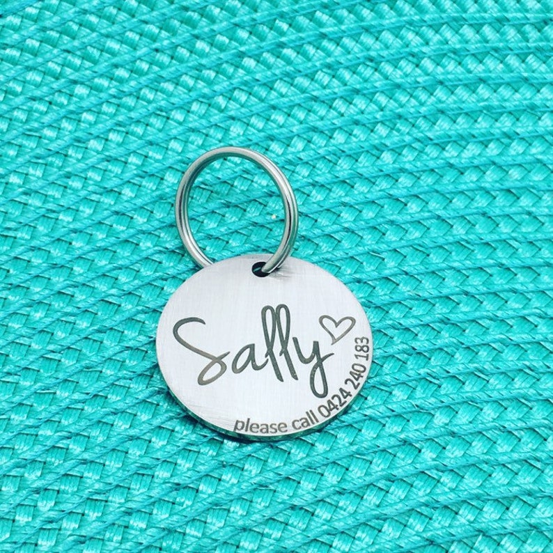 May include: A round, silver pet tag with the name "Sally" in cursive, a heart symbol, and a phone number. The tag is attached to a silver ring. The background is a textured teal surface.