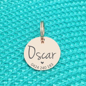K&ouml;nnte beinhalten: Ein ros&eacute;goldener Haustieranh&auml;nger mit Ring zur Befestigung. Der runde Anh&auml;nger ist mit dem Namen "Oscar", einem Herzsymbol und einer Telefonnummer graviert. Der Hintergrund ist eine t&uuml;rkisfarbene Textur.