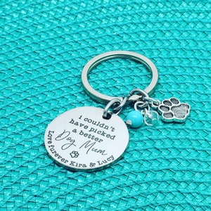 May include: A silver keychain with a round pendant engraved with "I couldn't have picked a better Dog Mum" and paw prints. It includes a paw print charm, a turquoise bead, and clear crystal accents. The text "Love furever Kira & Lucy" is also engraved.