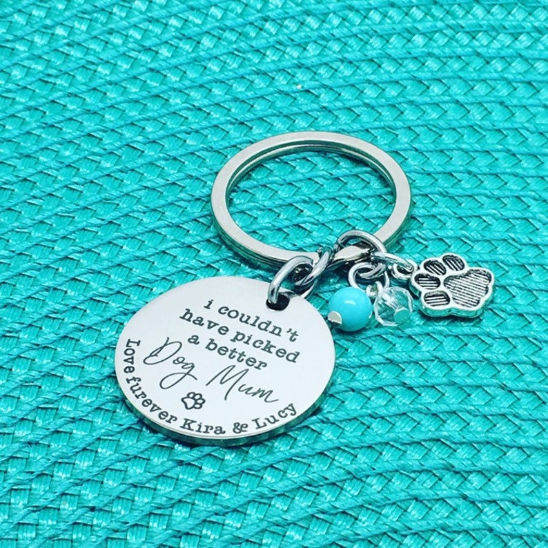 May include: A silver keychain with a round pendant engraved with "I couldn't have picked a better Dog Mum" and paw prints. It includes a paw print charm, a turquoise bead, and clear crystal accents. The text "Love furever Kira & Lucy" is also engraved.