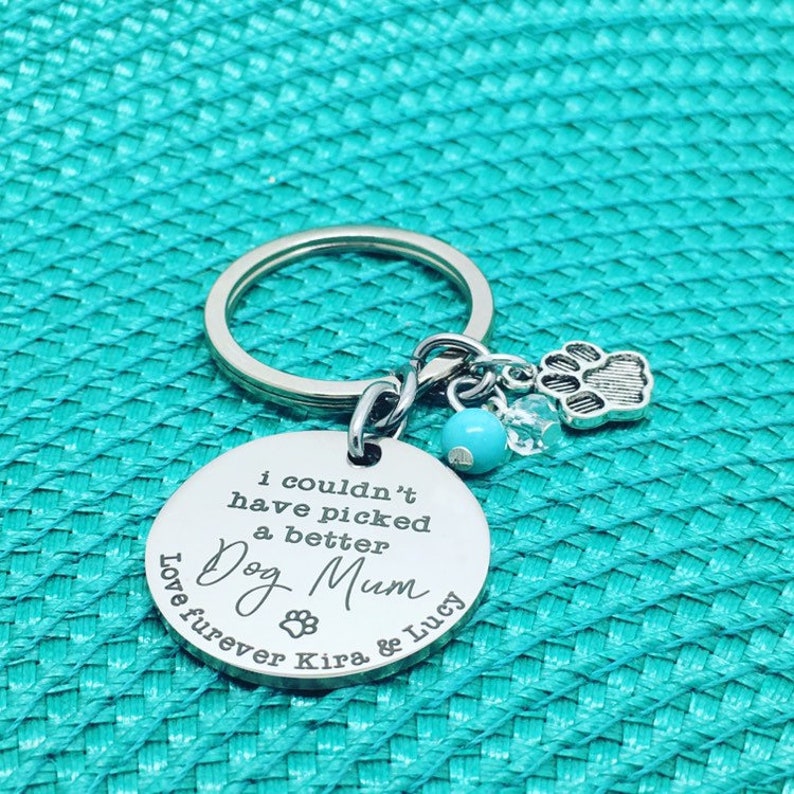 May include: A silver keychain with a round pendant that reads "I couldn't have picked a better Dog Mum" with a paw print and the names Kira & Lucy. The keychain has a paw print charm, a blue bead, and clear crystals.