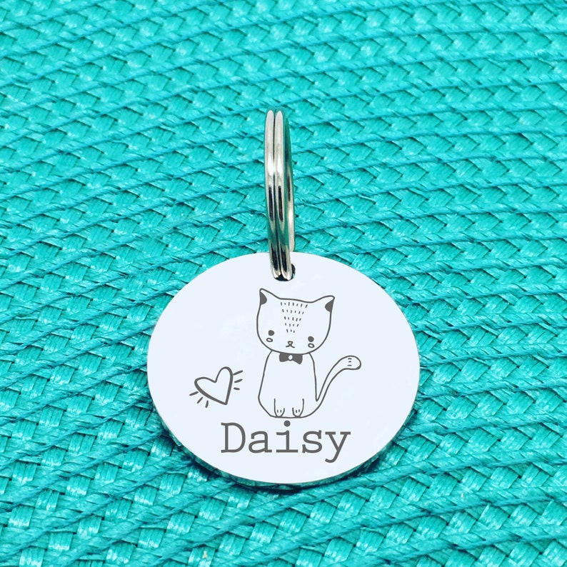 May include: A round, silver pet tag with a cartoon cat illustration and the name "Daisy" engraved below. The cat has a bow tie and a heart symbol next to it. A silver ring is attached at the top for easy attachment to a collar.