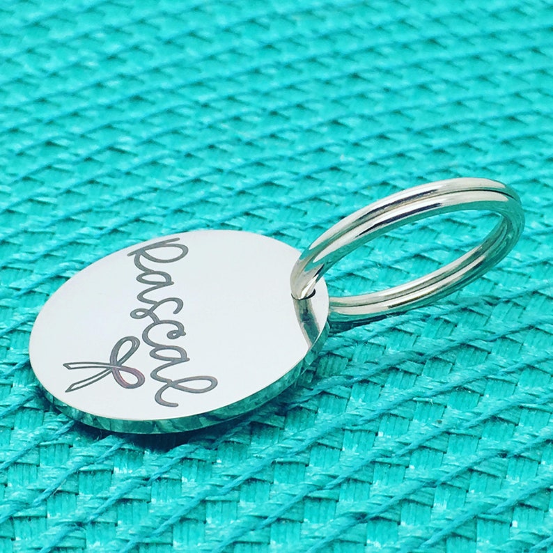 May include: A silver-colored oval keychain with the name "Pascal" in cursive script and a pink ribbon design. The keychain has a split ring for attaching keys. The background is a textured turquoise surface.