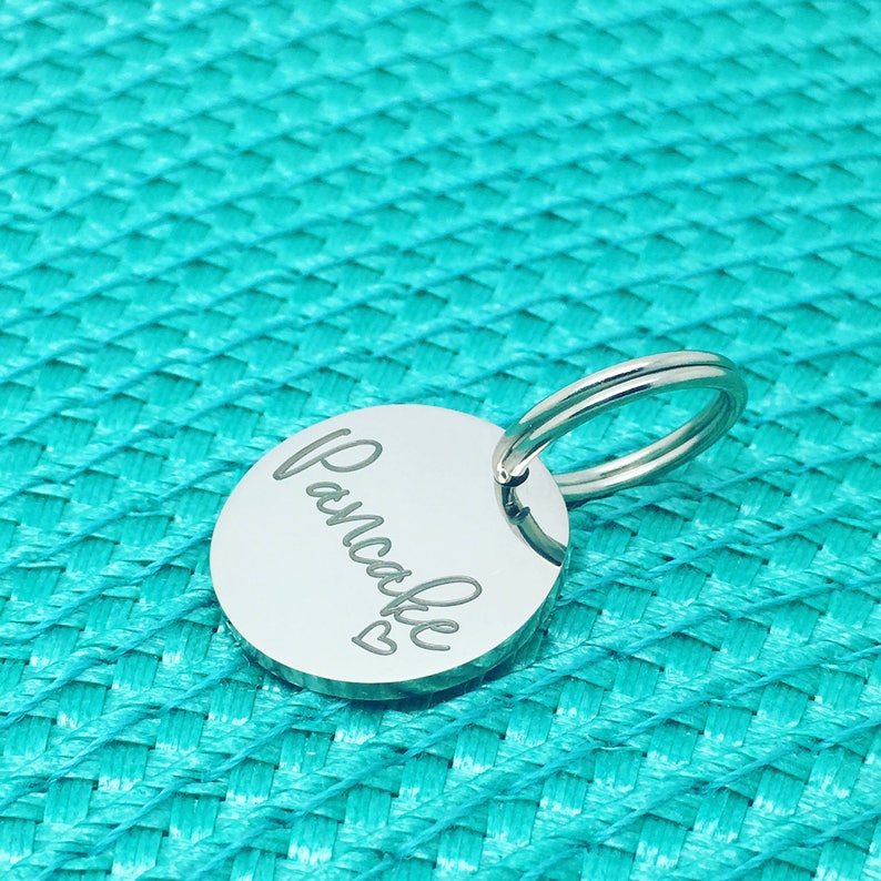 May include: A silver keychain with a round tag that says "Pancake" with a small heart symbol. The keychain is on a turquoise textured background.