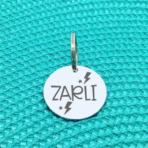 May include: A round, silver pet tag with the name "ZARLI" in a bold, sans-serif font. The tag features lightning bolt and star graphics. A silver split ring is attached at the top for easy attachment to a collar. The tag is set against a textured, turquoise background.