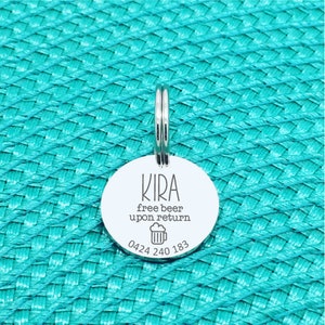 Puede incluir: Una placa redonda y plateada para mascotas con el nombre "KIRA" y la frase "free beer upon return" en texto negro. Un icono de jarra de cerveza y un n&uacute;mero de tel&eacute;fono tambi&eacute;n est&aacute;n impresos en la placa. La placa est&aacute; unida a un anillo plateado.