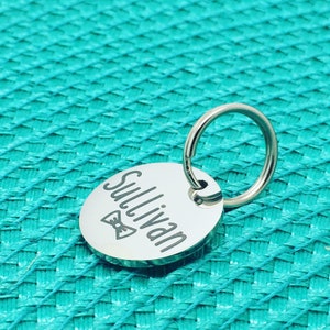 May include: A silver-colored, oval pet tag with the name "Sullivan" engraved in a script font. A small bow tie graphic is below the name. The tag is attached to a silver split ring. The background is a textured teal surface.