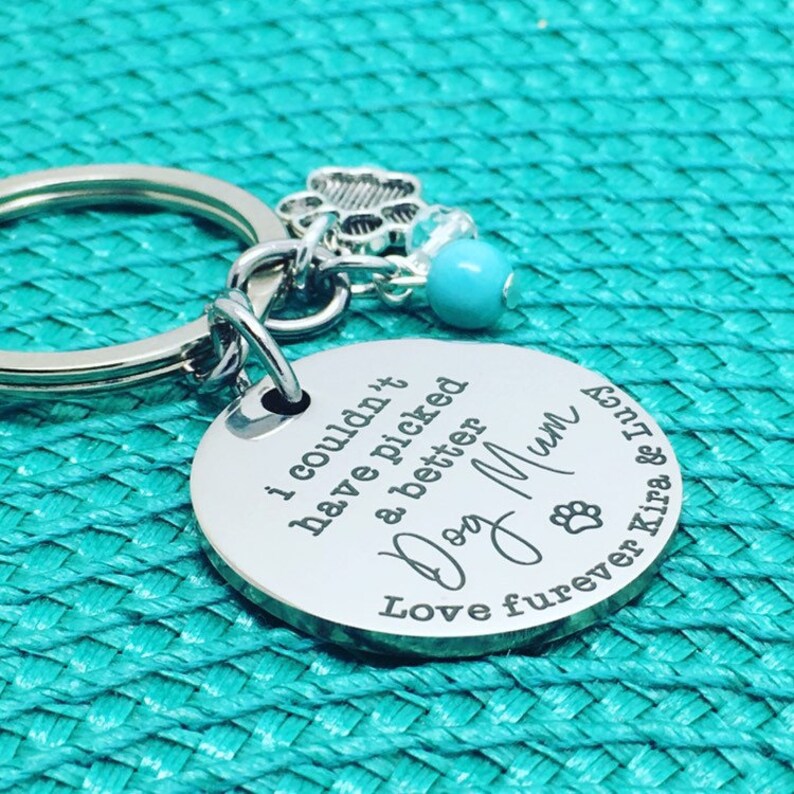 May include: A silver-colored keychain with a round pendant engraved with the words "I couldn't have picked a better Dog Mum". The keychain includes a paw print charm, a turquoise bead, and the names Kira & Lucy.