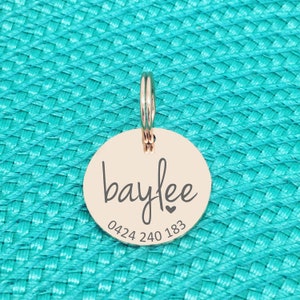 K&ouml;nnte beinhalten: Ein runder, ros&eacute;goldener Haustieranh&auml;nger mit dem Namen "baylee" in Schreibschrift, einem kleinen Herz und einer Telefonnummer. Der Anh&auml;nger ist an einem Metallring befestigt. Der Hintergrund ist eine strukturierte t&uuml;rkisfarbene Oberfl&auml;che.