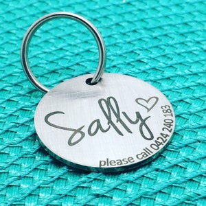 May include: A round, silver pet tag with a key ring. The tag is engraved with the name "Sally" in a cursive font, a heart symbol, and the phone number 0424 240 183. The words "please call" are also engraved on the tag.