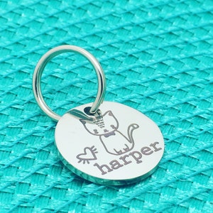 May include: A silver-colored, round pet tag with a cat illustration and the name "harper" engraved. The tag includes a split ring for attachment. A heart graphic is next to the cat. The tag is on a turquoise background.
