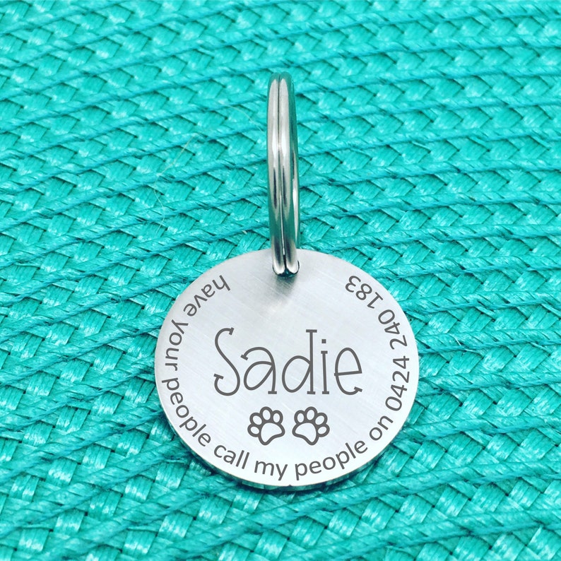 May include: A round, silver pet tag with the name "Sadie" engraved in a cursive font, along with paw prints. The tag also includes the phrase "Have your people call my people on 0424 240 183". A silver ring is attached at the top.
