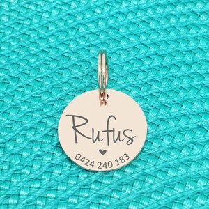 K&ouml;nnte beinhalten: Ein ros&eacute;goldener Haustieranh&auml;nger mit dem Namen "Rufus" in Schreibschrift, einem kleinen Herz und einer Telefonnummer. Der Anh&auml;nger ist rund und an einem silbernen Ring befestigt. Der Hintergrund ist eine strukturierte t&uuml;rkisfarbene Oberfl&auml;che.