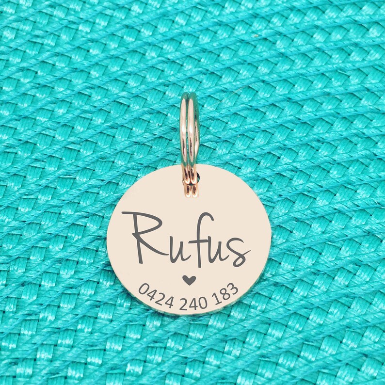 K&ouml;nnte beinhalten: Ein ros&eacute;goldener Haustieranh&auml;nger mit dem Namen "Rufus" in Schreibschrift, einem kleinen Herz und einer Telefonnummer. Der Anh&auml;nger ist rund und an einem silbernen Ring befestigt. Der Hintergrund ist eine strukturierte t&uuml;rkisfarbene Oberfl&auml;che.