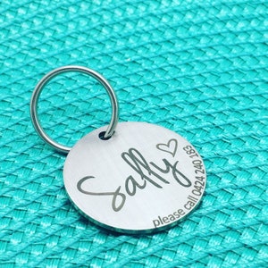 May include: A round, silver pet tag with the name "Sally" in cursive, a heart symbol, and a phone number. The tag is attached to a silver keyring. The background is a textured teal surface.