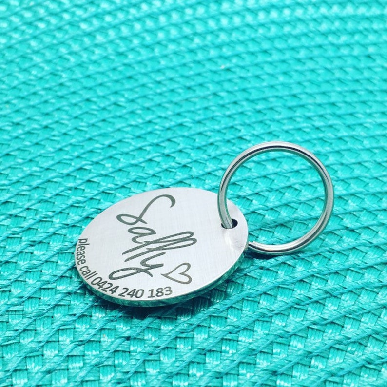 May include: Oval-shaped, silver-colored pet tag with the name "Sally" engraved in a cursive font, accompanied by a heart symbol. The tag includes the phone number 0424 240 183. A silver ring is attached for easy attachment to a collar.