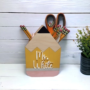 May include: A yellow and pink pencil-shaped desk organizer with the name "Ms. White" on the front. The organizer has a black eraser on top and holds pencils and scissors.