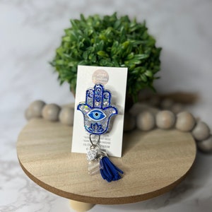 May include: A blue and silver glitter Hamsa hand charm with a blue evil eye. The charm is attached to a blue tassel and a clear acrylic charm with rhinestones. The charm is on a white card with the text "Jaya Creations" and "Follow us on Instagram @jayacreations".