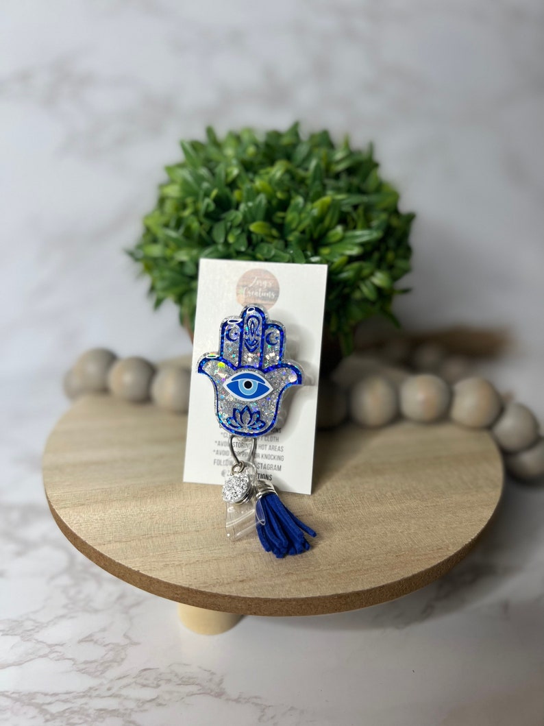 May include: A blue and silver glitter Hamsa hand charm with a blue evil eye. The charm is attached to a blue tassel and a clear acrylic charm with rhinestones. The charm is on a white card with the text "Jaya Creations" and "Follow us on Instagram @jayacreations".