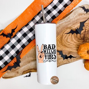 May include: White stainless steel tumbler with a black and white graphic that says "Bad Witch Vibes" and a peace sign with black nail polish. The tumbler is on a wooden surface with orange and black checkered ribbon and black bats.