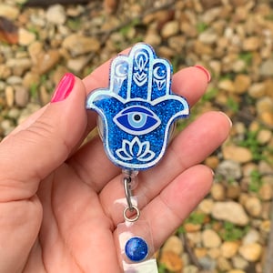 May include: Blue glitter Hamsa hand charm with a white evil eye and a white lotus flower design. The charm is attached to a clear plastic retractable badge holder with a blue glitter button.
