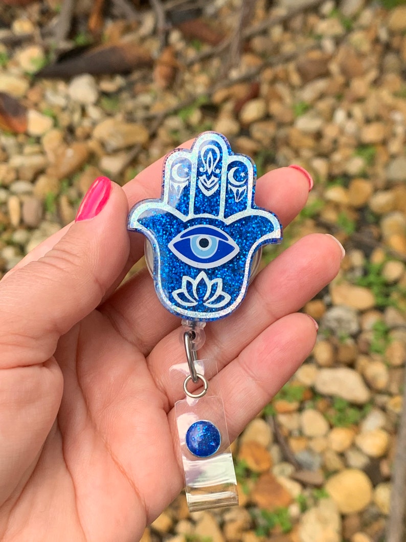May include: Blue glitter Hamsa hand charm with a white evil eye and a white lotus flower design. The charm is attached to a clear plastic retractable badge holder with a blue glitter button.
