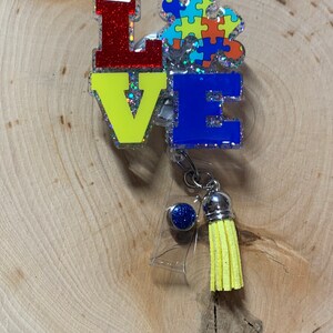 May include: A colorful "LOVE" badge with a puzzle piece design in blue, yellow, red, and orange. The badge has a clear plastic backing and a yellow tassel.