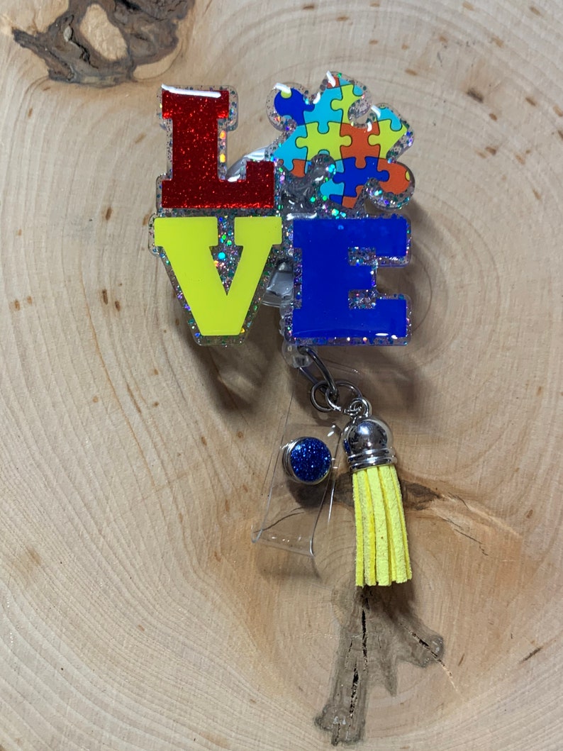 May include: A colorful "LOVE" badge with a puzzle piece design in blue, yellow, red, and orange. The badge has a clear plastic backing and a yellow tassel.