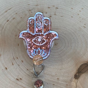 May include: A pink and gold glitter resin hamsa hand charm with a blue eye design. The charm is attached to a clear retractable badge holder with a silver clip and a small round pink and gold glitter charm.