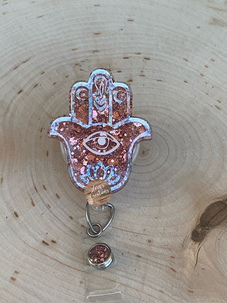 May include: A pink and gold glitter resin hamsa hand charm with a blue eye design. The charm is attached to a clear retractable badge holder with a silver clip and a small round pink and gold glitter charm.