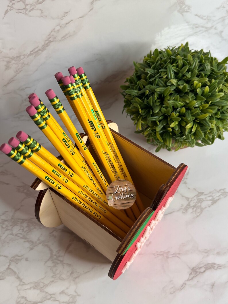 May include: A wooden watermelon pencil holder with a pink and green watermelon design. The holder is filled with yellow pencils with green and red accents. The pencils are labeled "Dixon Ticonderoga Soft".