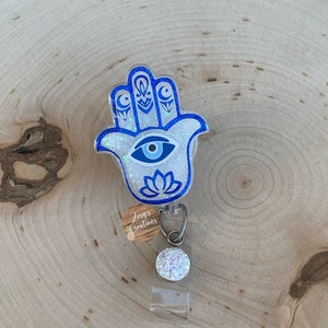 May include: White glitter Hamsa hand charm with a blue evil eye and blue crescent moons. The charm is attached to a clear plastic badge holder with a white glitter button.