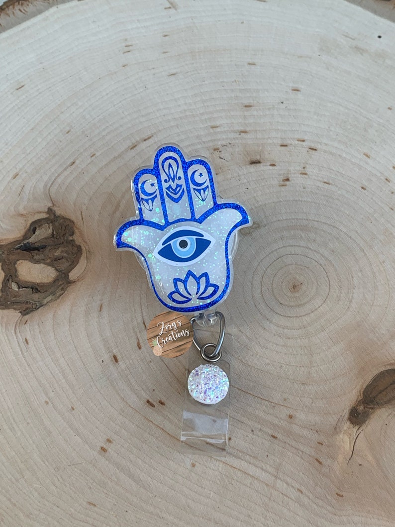 May include: White glitter Hamsa hand charm with a blue evil eye and blue crescent moons. The charm is attached to a clear plastic badge holder with a white glitter button.