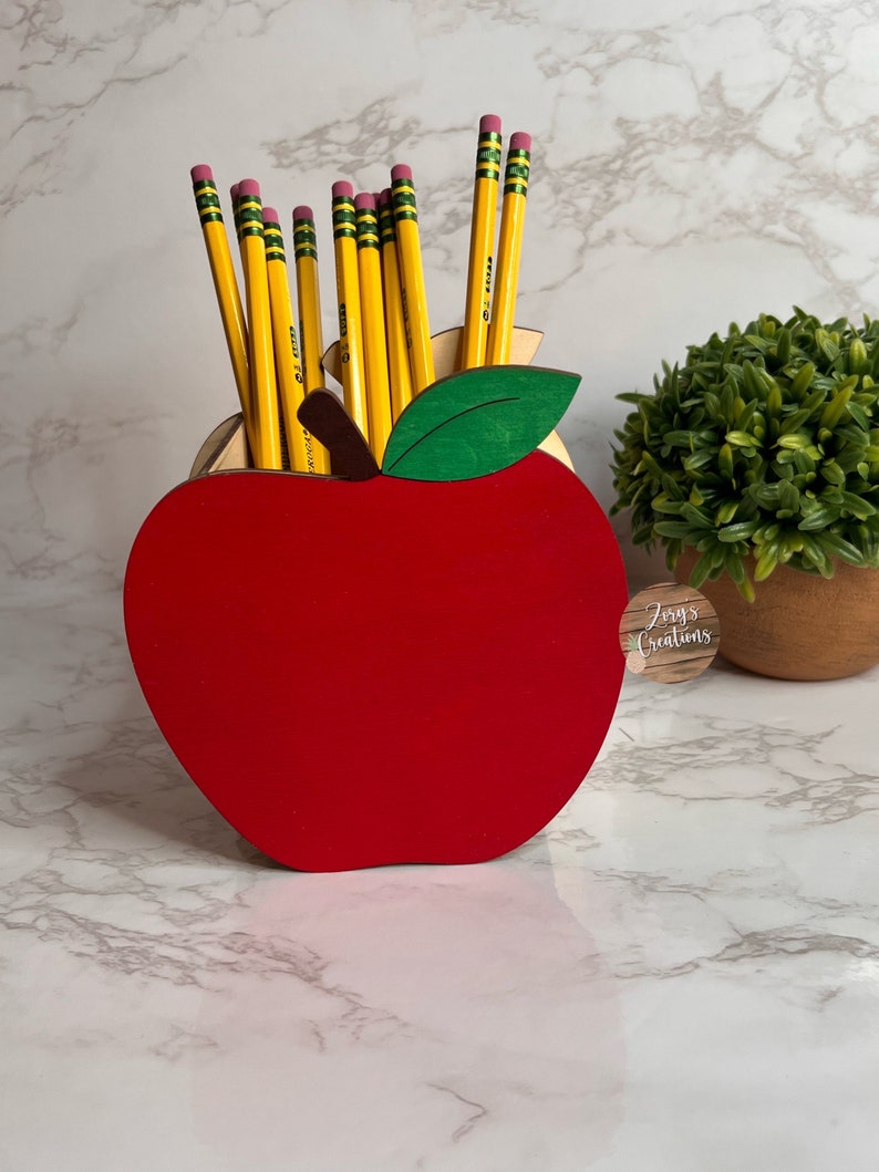 May include: A red wooden apple pencil holder with a green leaf and a brown wooden base. The holder is filled with yellow pencils with pink erasers. The holder is on a white marble surface.