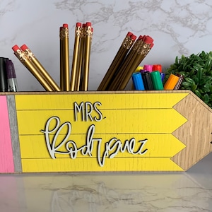 May include: A yellow pencil-shaped wooden desk organizer with a pink eraser, a gray stripe, and the text "MRS. Rodriguez" in white. The organizer is filled with gold pencils and colorful markers.
