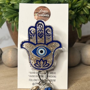 May include: A blue and gold glitter Hamsa hand charm with a blue evil eye in the center. The charm is attached to a clear plastic badge holder with a blue tassel. The text "Zory's Creations" is visible on the white card behind the charm.