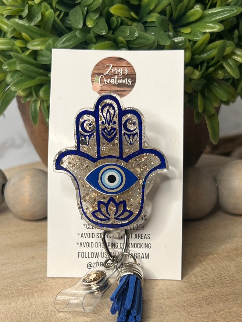 May include: A blue and gold glitter Hamsa hand charm with a blue evil eye in the center. The charm is attached to a clear plastic badge holder with a blue tassel. The text "Zory's Creations" is visible on the white card behind the charm.
