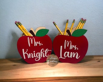 May include: Two red wooden apple pencil holders with green leaves. The holders are personalized with the names "Mrs. Knight" and "Mrs. Lam" in white lettering.