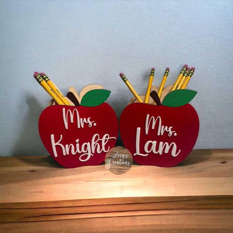 May include: Two red wooden apple pencil holders with green leaves. The holders are personalized with the names "Mrs. Knight" and "Mrs. Lam" in white lettering.