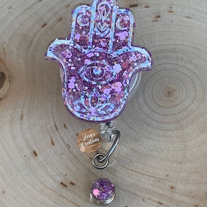 May include: Purple glitter Hamsa hand charm with a blue eye and a silver retractable badge reel. The charm is attached to a clear plastic badge holder with a silver clip. The badge holder has a small, round, purple glitter charm attached to it.