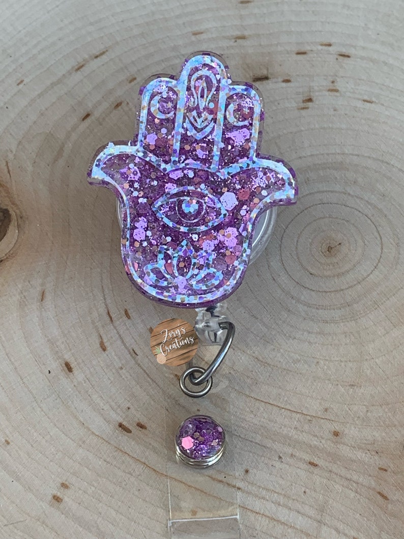 May include: Purple glitter Hamsa hand charm with a blue eye and a silver retractable badge reel. The charm is attached to a clear plastic badge holder with a silver clip. The badge holder has a small, round, purple glitter charm attached to it.