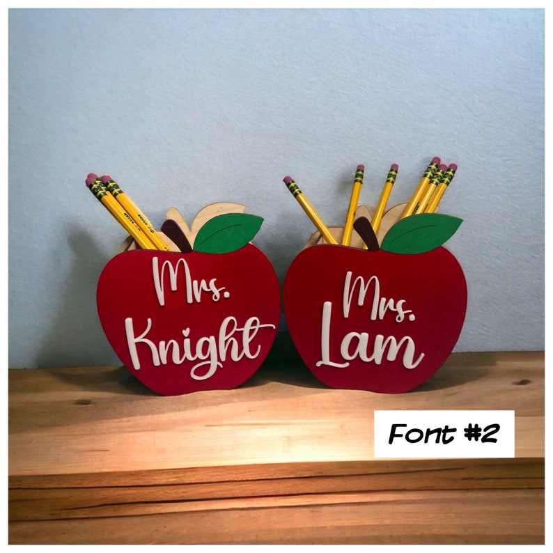 May include: Two red wooden apple pencil holders with green leaves. The holders are personalized with the names "Mrs. Knight" and "Mrs. Lam" in white lettering. The text "Font #2" is visible below the holders.