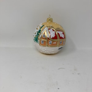 Op de afbeelding: Een ronde, gouden kerstbal met een wit en rood geschilderd huis met sneeuw op het dak. De bal heeft een witte achtergrond met groene bomen en witte sneeuwvlokken.