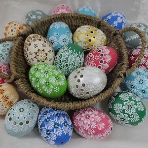 Pode incluir: Uma cesta de vime cheia de ovos de Páscoa decorados de forma intrincada. Os ovos são pintados em várias cores, incluindo verde, azul, rosa e branco, com desenhos florais intrincados.