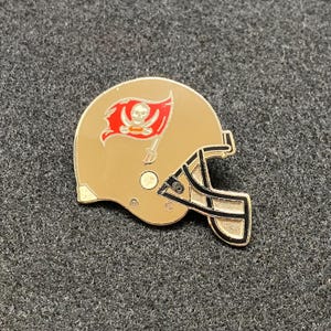 May include: A gold football helmet pin with a red pirate flag and skull and crossbones design. The helmet is black and gold.