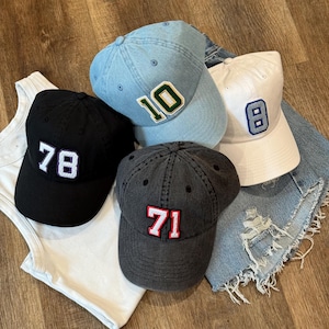 May include: Four baseball caps in black, light blue, white, and charcoal gray. Each cap features a different number: 78, 10, 8, and 71. The numbers are in white or blue with a contrasting border. The caps are on a wooden surface.