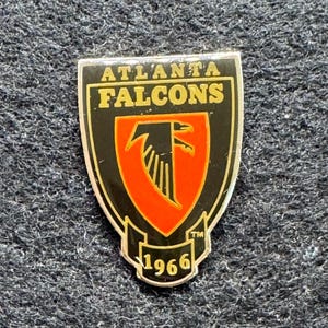 May include: A shield-shaped Atlanta Falcons lapel pin featuring a black background with gold lettering. The center displays a red shield with a black falcon logo. A small banner at the bottom contains the gold text 1966 and a trademark symbol.