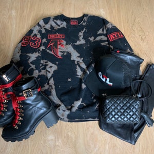 Może przedstawiać: Czarno-szara bluza w stylu tie-dye z czerwonymi haftowanymi naszywkami z napisami "93", "Bailey" i "ATL". Bluza leży na drewnianej podłodze z czarnymi skórzanymi spodniami, czarną pikowaną torebką i czarnymi butami z czerwonymi sznurowadłami.