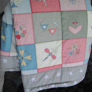 Könnte beinhalten: Eine Patchwork-Decke mit verschiedenen bunten Designs, darunter Vögel, Blumen und Herzen. Die Decke ist aus weichem Baumwollstoff gefertigt und hat eine weiße Rückseite mit Sternen.